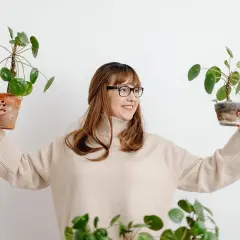 Mujer sonriente con gafas sostiene dos plantas en maceta, mostrando flores que no necesitan sol.