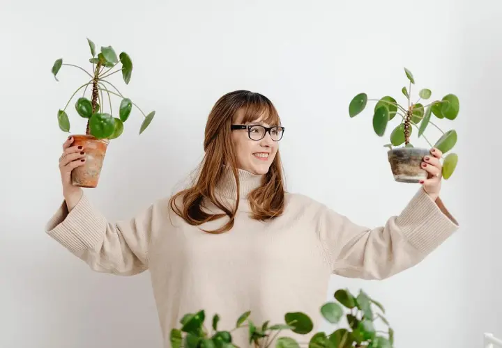 Mujer sonriente con gafas sostiene dos plantas en maceta, mostrando flores que no necesitan sol.