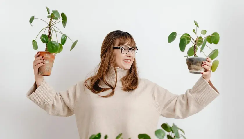 Mujer sonriente con gafas sostiene dos plantas en maceta, mostrando flores que no necesitan sol.