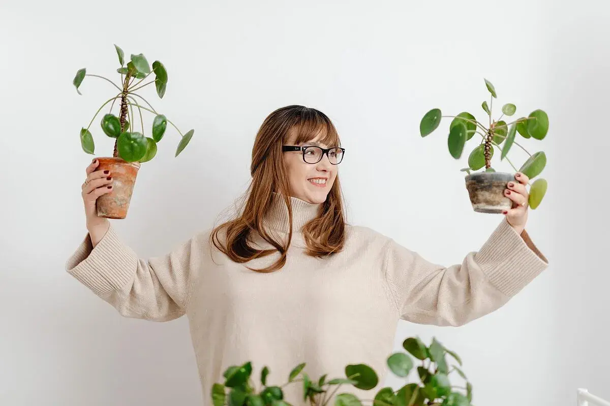 Mujer sonriente con gafas sostiene dos plantas en maceta, mostrando flores que no necesitan sol.