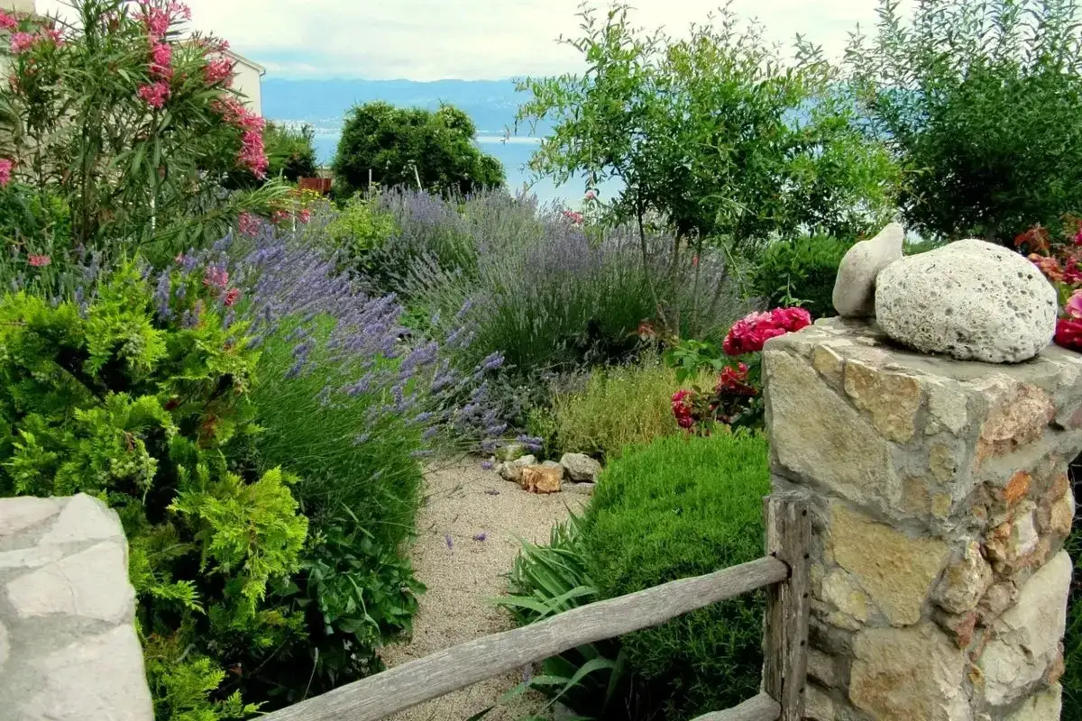 Jardín con lavanda, rosas y exuberantes flores verdes. Un camino de grava serpentea entre la vegetación, con el mar y las montañas al fondo.